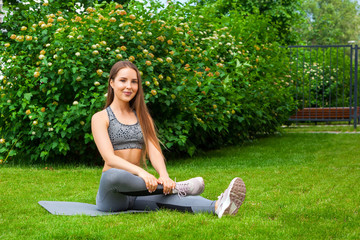 Beautiful thin woman sportswoman doing the exercise, stretches legs before training on a yoga mat on a green lawn in the park on a summer day