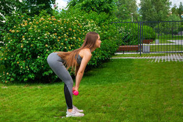 Fototapeta premium A dark-haired woman coach in a sporty short top and gym leggings makes a deadlift with dumbbells on a summer day in a park on a green lawn