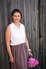 Beautiful dark-haired woman in white shirt and beige skirt smiling, posing and holding in her hand a big bouquet of pink peonies on the background of wooden walls...