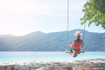 Outdoors lifestyle fashion portrait stunning young girl enjoying on swing on the tropical island. In the background the sea. Wearing stylish dress.