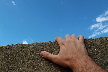 Adult male man hand holding edge of small concrete shed roof, trying to climb on it. Blue skies in background. First person view is recreating feel of FPS or survival computer games or extreme sports