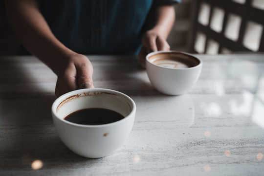 Closeup image of hands holding two white cups of hot coffee on table in cafe