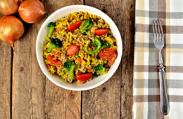 View from above of bowl with tuna risotto with vegetables, tomatoes, broccoli and parsley, onions and brown checkered tablecloth in right frame