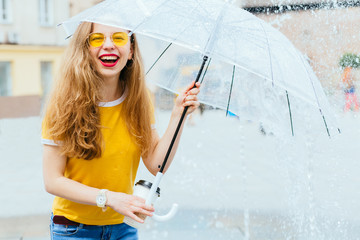 Portrait of hipster excited girl in yellow eyeglesses breathing fresh air with umbrella plaing with fountains water jets in city squre urban background. back to childhood concept.