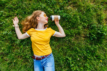 Relaxed hipster woman in yellow t-shirt with cup of coffe lying on the grass. Summer energy drink morning concept.