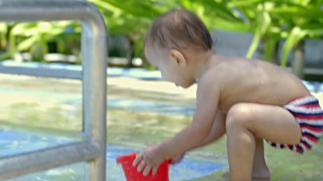 Cute toddler boy playing with toy plastic bucket in shallow water at the edge of kiddie swimming pool at hot summer day