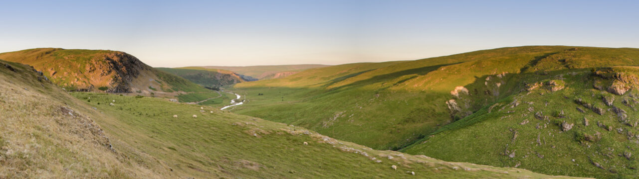 Panoramic view of the spectacular Elan Valley, in Powys, mid Wales. Sheep are grazing in the landscape and a small stream runs through the valley. The setting sun is lighting the mountain tops