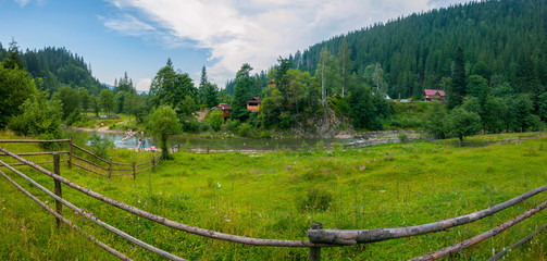 Fototapeta premium Beautiful views of the mountainous terrain with the current river between the low banks with green grass on one side and trees and houses on the other.