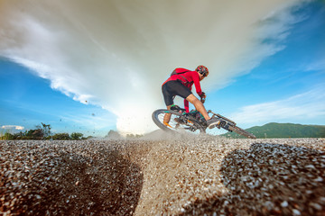 Low, wide angle portrait against explose blue sky of mountain biker going downhill. Cyclist in red sport equipment and helmet slice bike through the ground at risk of accident