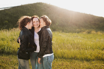 Concept of youth and maturity. Nice mother hugs her adult daughters dressed in the same clothes with a white T-shirt in jeans and leather jackets against the backdrop of a green hill on a warm sunny d