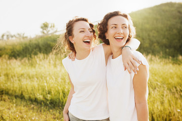 Portrait of cute young beautiful curly-haired girls sisters standing into the distance dressed in a white T-shirt posing against a green hill - obrazy, fototapety, plakaty