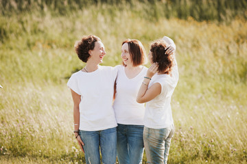 Family portrait of a positive mother hugs her daughters in white T-shirts and jeans against the backdrop of a green hill on a summer sunny day.