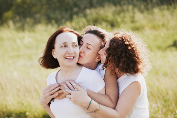 Three pretty women mum and two adult daughters embrace while walking outside the city on a background of a blurred picturesque hill