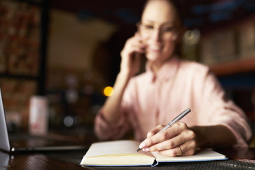 Young woman freelancer writing with pen ideas in notepad while using mobile phone. focus on woman s...