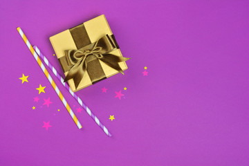 Golden shiny classic gift box with brown satin bow and cocktail straws with confetti in the shape of stars as attributes of party