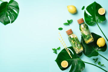 Bottle of detox water with mint, lemon and tropical monstera leaves on blue background. Flat lay. Citrus lemonade. Summer fruit infused water. Top view with copy space.
