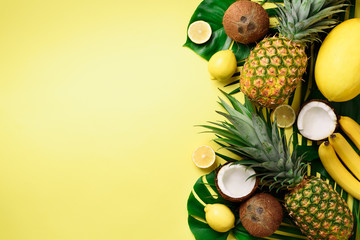 Exotic pineapples, ripe coconuts, banana, melon, lemon, tropical palm and green monstera leaves on yellow background with copyspace. Creative layout. Monochrome summer concept. Flat lay, top view.