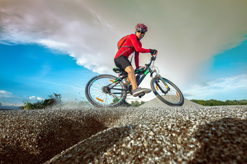 Naklejka premium Low, wide angle portrait against explose blue sky of mountain biker going downhill. Cyclist in red sport equipment and helmet slice bike through the ground at risk of accident