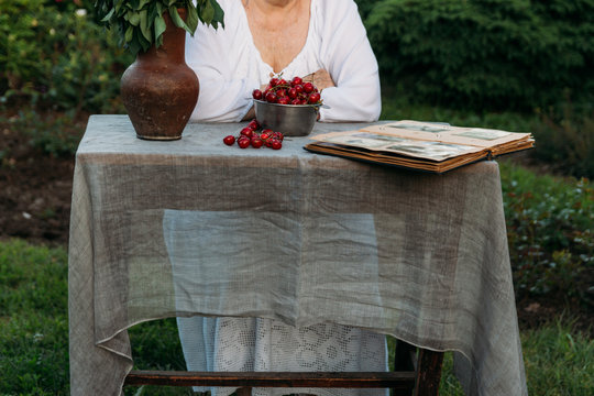 grandmother sitting in a chair at the table on which the cherry is lying, in the garden and watching an old album with photos, recounts and remembers the family's story. grandmother in a white vintage