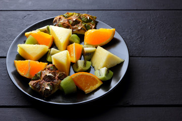 Fruit salad on a black plate on wooden boards, pineapples, oranges and kiwi on a dark background, salad of chopped tropical fruits for breakfast, vegetarian food, copy space