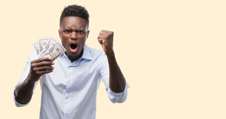 Young african american businessman holding dollars annoyed and frustrated shouting with anger, crazy and yelling with raised hand, anger concept