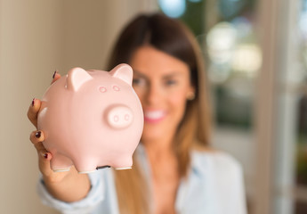 Close up of beautiful young woman holding piggy bank at home, very happy with a smile because of...