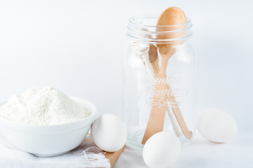  Baking ingredients. White background. Ingredients and tools to make a cake. Flour, milk, eggs, rolling pin, wooden spoons, jar. Copy space. Bakery. Baking concept