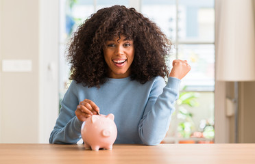 Obraz na płótnie Canvas African american woman saves money in piggy bank screaming proud and celebrating victory and success very excited, cheering emotion