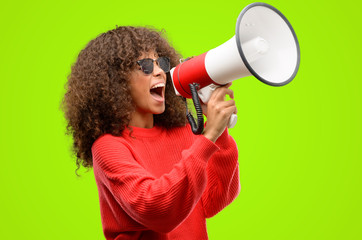 African american woman wearing sunglasses communicates shouting loud holding a megaphone,...