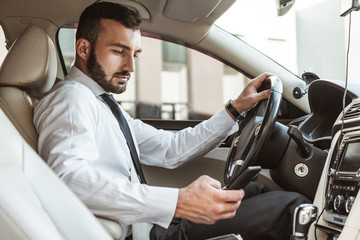 handsome driver in shirt driving car and looking at smartphone
