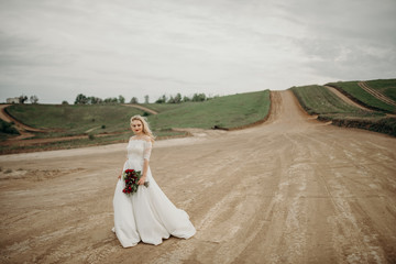 Fototapeta na wymiar The joyful stylish beautiful fabulous happy blonde bride with the stylish bouquet is tenderly hugging flowers on the road in the countryside during the cloudy sunset.