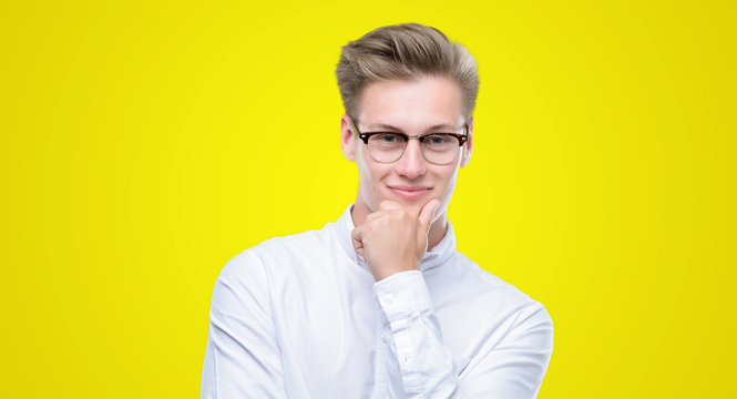 Young handsome blond man looking confident at the camera with smile with crossed arms and hand raised on chin. Thinking positive.