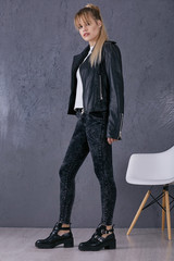 Full length isolated studio portrait of a young blonde lady in a white T-shirt, black tie-dye jeans, a leather biker jacket and ankle boots. The side view of the girl, standing, looking at the camera.