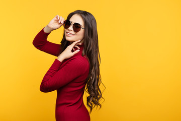 Pretty young lady in red dress and sun glasees is posing with hand on face on orange background.