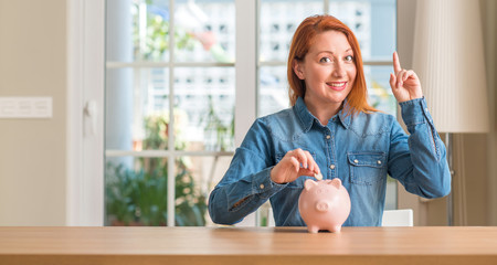 Redhead woman saves money in piggy bank at home surprised with an idea or question pointing finger...
