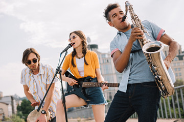 Multiracial young people with guitar, djembe and saxophone performing on street - obrazy, fototapety, plakaty