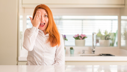 Redhead woman at kitchen covering one eye with hand with confident smile on face and surprise emotion.