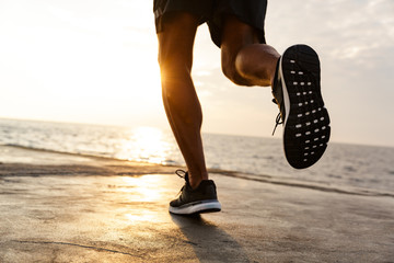 Cropped male legs of healthy sportsman wearing shorts and sneakers, running along pier at seaside...