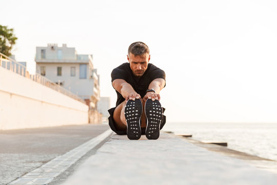Image of caucasian sportsman 30s in tracksuit sitting on parapet, and stretching legs near seaside during summer morning