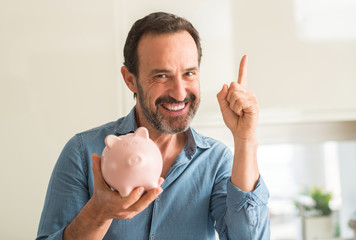 Middle age man save money on piggy bank surprised with an idea or question pointing finger with...