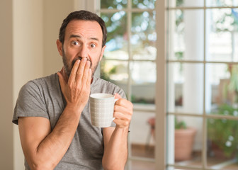 Middle age man drinking coffee in a cup cover mouth with hand shocked with shame for mistake, expression of fear, scared in silence, secret concept