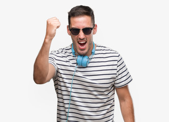 Naklejka premium Handsome young man wearing headphones angry and mad raising fist frustrated and furious while shouting with anger. Rage and aggressive concept.