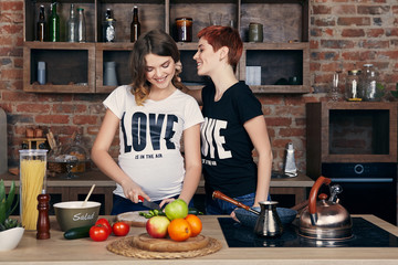 Lesbian pregnancy. Two young healthy women, one of them pregnant, enjoying togetherness in the kitchen. The beautiful expectant mom cutting vegetables while her girlfriend touching her hair tenderly.