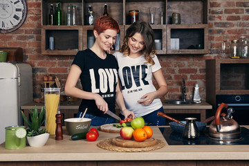 Lesbian pregnancy. Two happy young women, one of them pregnant, enjoying togetherness in the kitchen. The expectant mom leaning on the shoulder of her girlfriend who's cutting cucumbers. Both smiling.