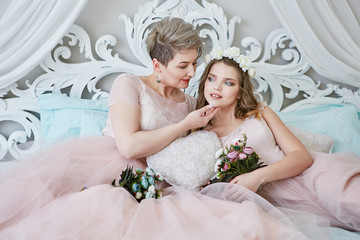 Lesbian honeymoon. Newlyweds in light pink bridal dresses lying on a luxury bed and enjoying togetherness, with bouquets in their hands. The short-haired bride touching tenderly her spouse's chin.