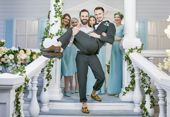 Gay wedding. One groom carrying the other down the steps of the front porch, embellished with...