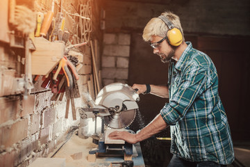 Young hipster bearded man with ear protectors by profession carpenter builder saws with a circular...