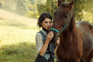 Obraz premium A young lady in a vintage dress, with tenderness and with affection hugs her horse. An ancient, collected hairstyle, a gentle make-up. Background of summer fields. Art photo