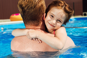 rear view of father embracing his little smiling daughter in swimming pool while she looking at camera