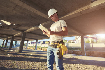 Smiling young man architect in building helmet standing and holding touch pad. Smiling construction manager standing on building site. Builder worker activity with digital tablet on construction site